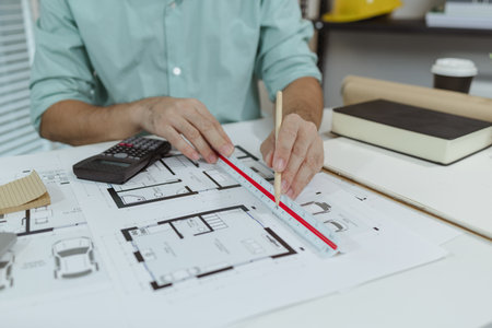 An architect working on house plans at a white desk with blueprints, a calculator, and a triangular...