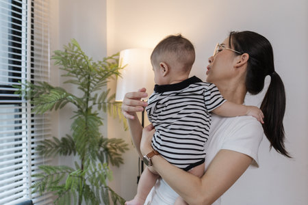A young mother is holding her baby while spending quality time together indoors. She looks at her child with love and care, highlighting the warmth and bond of family and parenthood.の写真素材