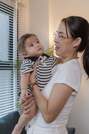 A young mother is holding her baby while spending quality time together indoors. She looks at her child with love and care, highlighting the warmth and bond of family and parenthood.の写真素材