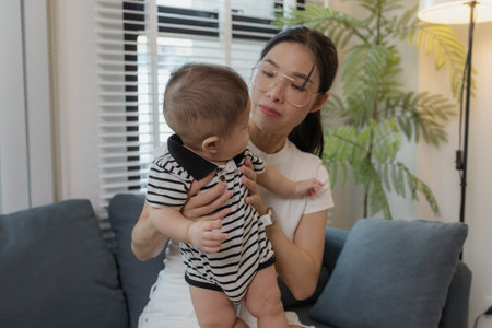 A young mother lovingly cradles her baby while breastfeeding on a sofa, symbolizing maternal care,...