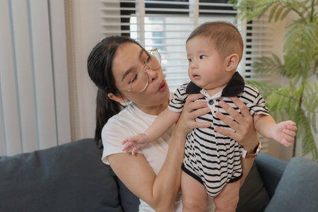 A young mother is holding her baby while spending quality time together indoors. She looks at her...