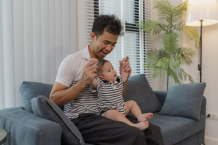 A cheerful father is holding his baby in his arms, sitting on the sofa at home. The baby looks...
