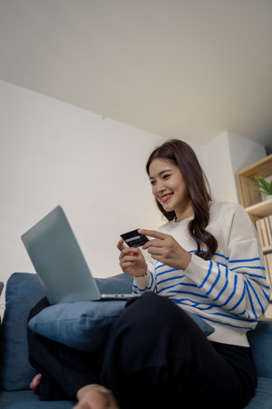 A young woman sits on a sofa using a laptop while holding credit cards, representing secure online payment and modern lifestyle. She smiles confidently while shopping from home.の写真素材