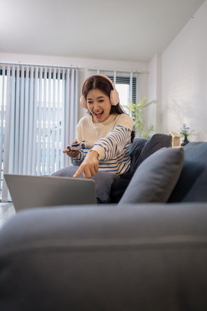 A young woman sits on a sofa wearing headphones while holding a game controller and playing a video game on her laptop. Her cheerful expression shows enjoyment and excitement.の写真素材