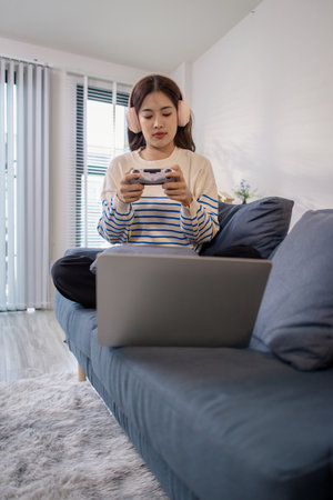 A young woman wearing headphones is playing games on her laptop using a joystick while sitting on the couch. She shows frustration and disappointment after losing the game.の写真素材