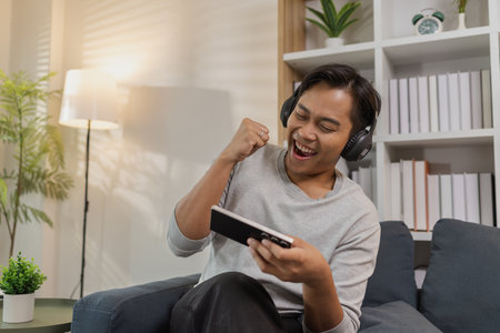 A young man is sitting comfortably on the sofa, wearing wireless headphones and playing a mobile...
