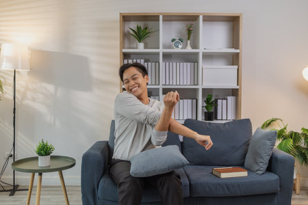 A young man in a gray shirt relaxes and stretches his arms comfortably while sitting on a cozy blue sofa at home. He enjoys a peaceful moment, smiling with contentment and feeling refreshed.の写真素材