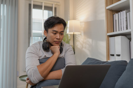 A young man is relaxing on a sofa at home, enjoying his leisure time while watching movies or streaming content on his laptop. The cozy modern living room creates a comfortable atmosphere.の写真素材