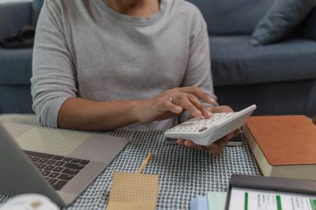 A young woman is working at home, using a laptop and calculator with a cheerful expression. She appears relaxed and focused, enjoying her remote work in a modern living room.の写真素材