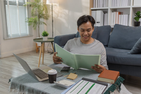 A young man is reviewing documents at home while working remotely. He looks focused and content, sitting in a modern living room surrounded by work materials and technology.の写真素材