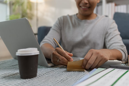 A young man is working from home, taking notes with a pencil while using his laptop. There is a cup of coffee on the table, and he appears focused in a cozy home office environment.の写真素材