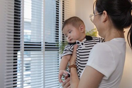 A young mother is holding her baby while spending quality time together indoors. She looks at her child with love and care, highlighting the warmth and bond of family and parenthood.の写真素材