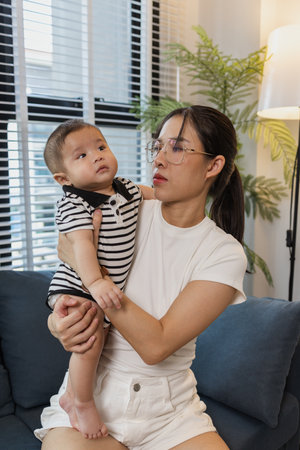 A young mother is holding her baby while spending quality time together indoors. She looks at her child with love and care, highlighting the warmth and bond of family and parenthood.の写真素材