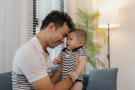 A cheerful father is holding his baby in his arms, sitting on the sofa at home. The baby looks...