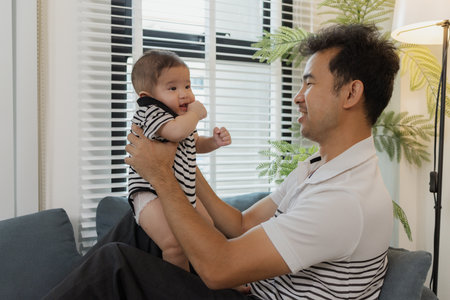 A tender moment of a father holding his baby, as the infant looks up with curiosity and trust. The...