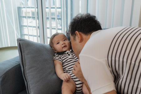 A cheerful father bonding with his baby in the living room, smiling warmly while holding the infant hands. The moment reflects love, joy, and the happiness of parenthood.の写真素材