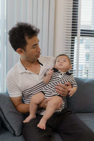 A cheerful father is holding his baby in his arms, sitting on the sofa at home. The baby looks...