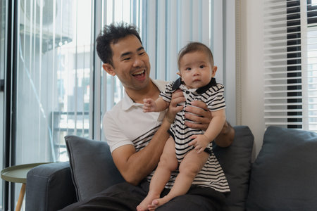 A cheerful father is holding his baby in his arms, sitting on the sofa at home. The baby looks...