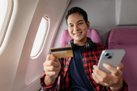 Cheerful young man sits on a window seat inside an airplane, holding a smartphone in one hand and a credit card in the other. He smiles, appearing to enjoy making an online purchase during his flight.の写真素材