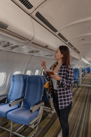 Young woman smiling and pointing while holding a passport and boarding pass inside an airplane cabin. She looks excited as she searches for her assigned seat at the start of her journey.の写真素材