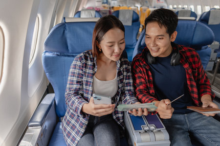A happy couple sitting together on an airplane, holding cash, credit cards, and smartphones. They appear to be managing their travel expenses or planning their spending during the flight.の写真素材