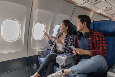 Young couple sitting together on an airplane, smiling and pointing out the window. They appear cheerful and engaged, enjoying the journey and spending meaningful travel time with one another.の写真素材