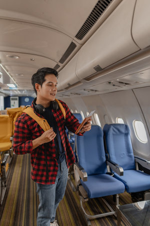 Asian man boarding an airplane and searching for his seat with a passport and boarding pass in hand. He looks happy and excited, carrying a backpack and wearing headphones around his neck.の写真素材