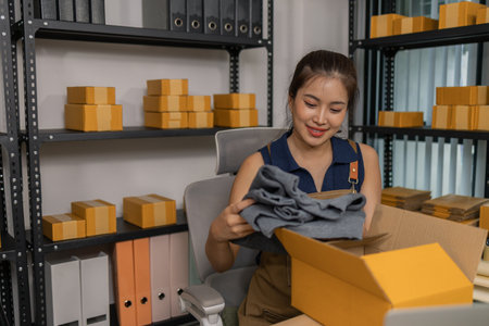 Smiling young female entrepreneur packing clothes into a parcel box, happily preparing merchandise...