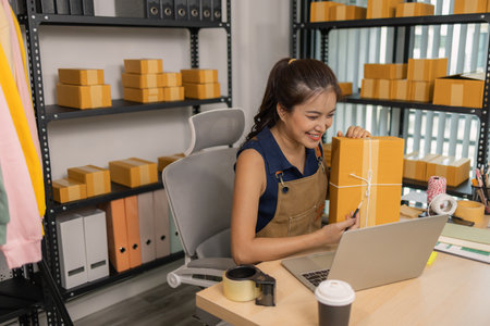 Young female entrepreneur smiling while holding a parcel and using a laptop at her home office. She manages an online store, prepares deliveries, and communicates with customers for e-commerce.の写真素材