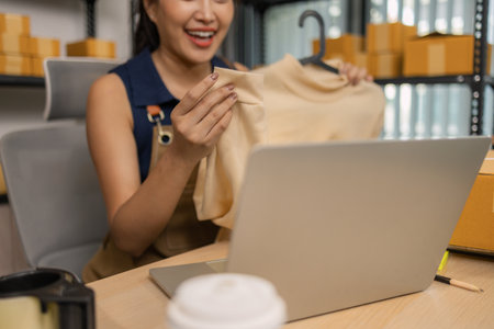 Young female entrepreneur smiling while livestreaming to sell clothes from her home office. She presents fashion items, interacts with viewers, and manages her online store during the live session.の写真素材