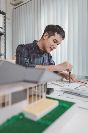 Young male architect working at office desk, drawing construction plans and using drafting tools,...