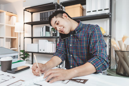 Young male architect working at office desk, drawing construction plans and using drafting tools,...