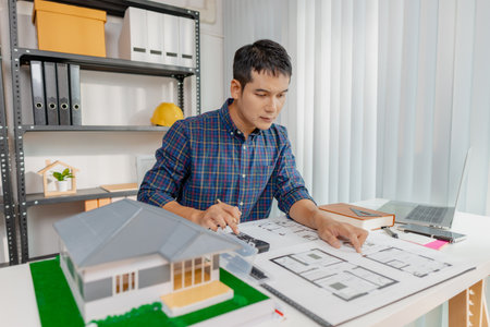 A young architect works on house design plans at his desk, focusing on drawings and scale models....