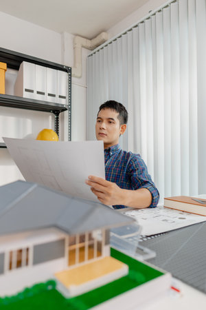 Young male architect working thoughtfully at office desk, analyzing house blueprints and...