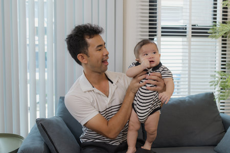 A cheerful father is holding his baby in his arms, sitting on the sofa at home. The baby looks...