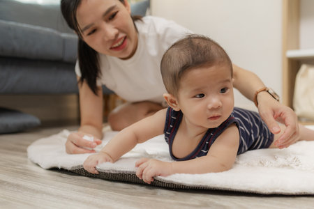 A baby enjoys tummy time on a soft mat while the mother stays close, encouraging him with a warm...