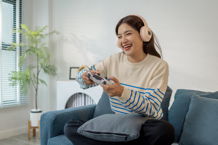 A young woman wearing headphones is sitting on a sofa while holding a game controller, showing joy,...