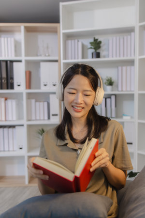 A young woman sits on a sofa with headphones, reading a book with a calm smile. She enjoys her...