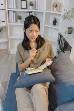 A young woman sits comfortably on a sofa in a modern home office, holding a notebook and writing thoughtfully. She appears focused and relaxed, enjoying a quiet moment of personal reflection.の写真素材