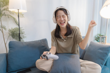 A young woman wearing headphones is sitting on a sofa with a game controller in her hand, smiling and raising her fist in celebration while playing a video game on her laptop.の写真素材