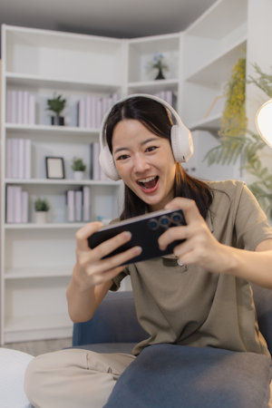 A young woman wearing headphones sits on a sofa, holding a smartphone horizontally with both hands, fully engaged in playing a mobile game, showing excitement and focus.の写真素材