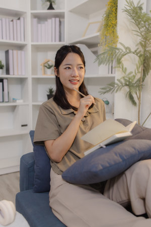 A young woman sits comfortably on a sofa in a modern home office, holding a notebook and writing...