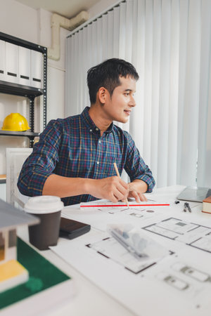 Young male architect working at office desk, drawing construction plans and using drafting tools,...