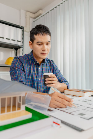 A young architect works on house design plans at his desk, focusing on drawings and scale models....
