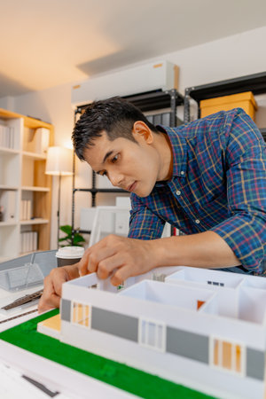 Young architect working in office with building models and floor plans, analyzing details and...