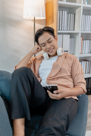 A young man relaxes at home, smiling while using his smartphone. He enjoys leisure time with...