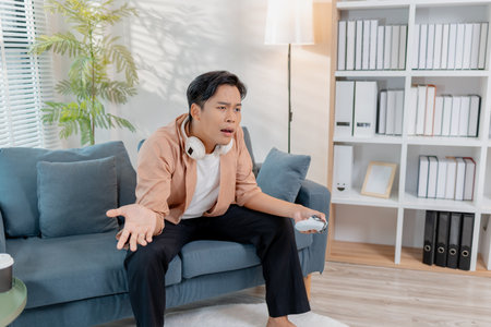 A young man sits on a sofa holding a game controller, fully focused and expressing frustration as he plays a video game. His intense reaction highlights the challenge and excitement of gaming.の写真素材