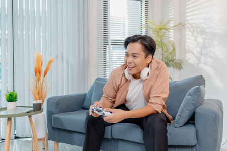 A cheerful young man is sitting on a sofa at home, holding a game controller and playing video games...