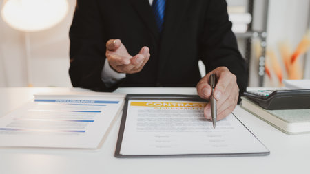 A businessman in a formal suit explains car insurance details, holding a small red car model and...