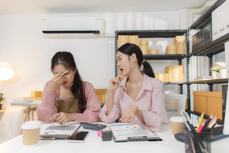 Two young women work together in a small business office, organizing packages, checking financial...